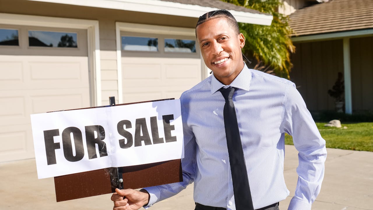 about-01 Real estate agent standing with a 'For Sale' sign in front of a house on a sunny day.